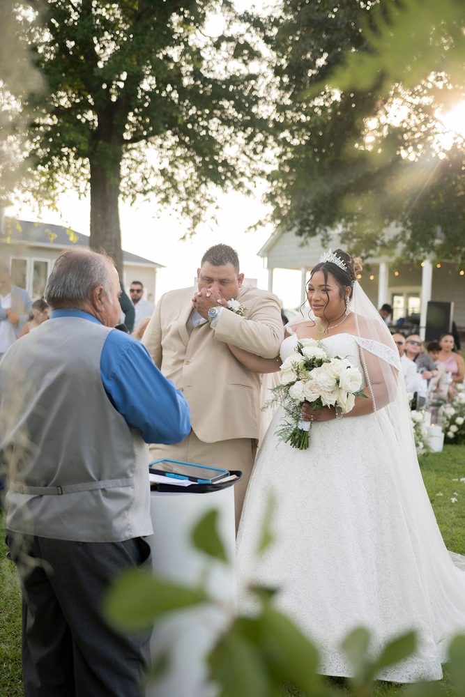 Emotional groom in a tan suit covering his mouth at the outdoor altar beside his bride in a tiara and lace gown