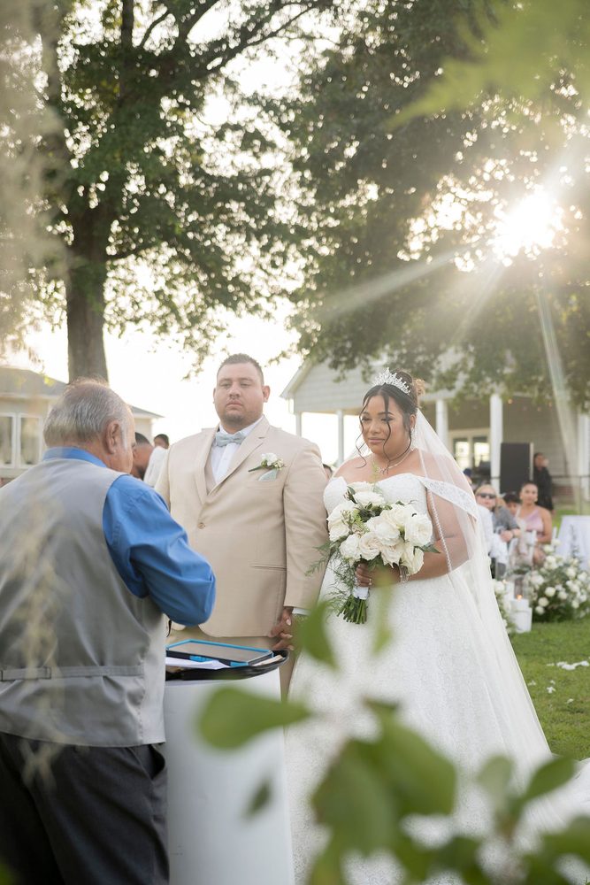 Bride in a tiara holding white roses beside the groom in a tan suit as the officiant reads, sun flaring through trees