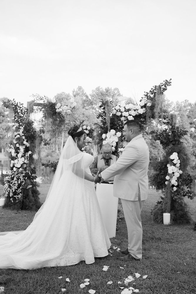 Black-and-white of the couple holding hands under a white-rose floral arch, petals scattered on the grass