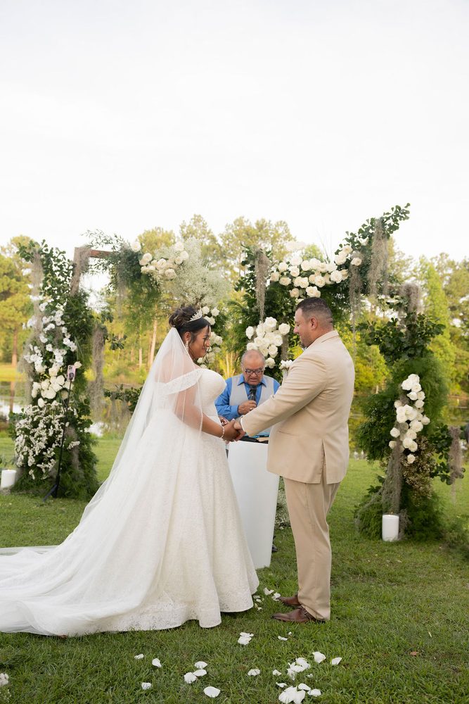 Bride and groom holding hands before the officiant under a white rose and moss arch by a pond
