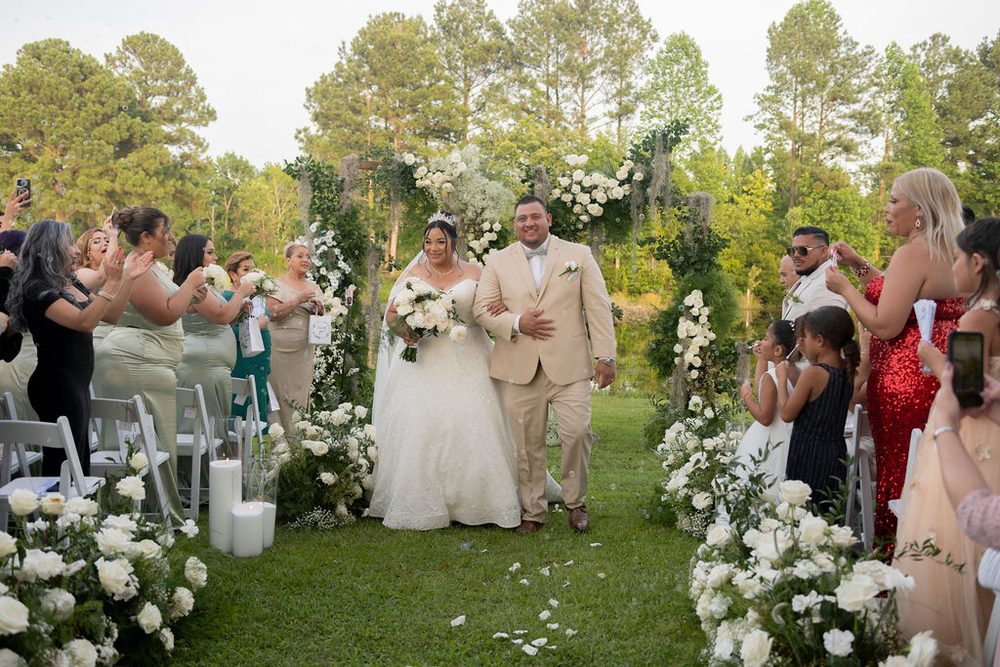 Newlyweds walking up a grassy aisle lined with white roses and candles as guests cheer and take photos