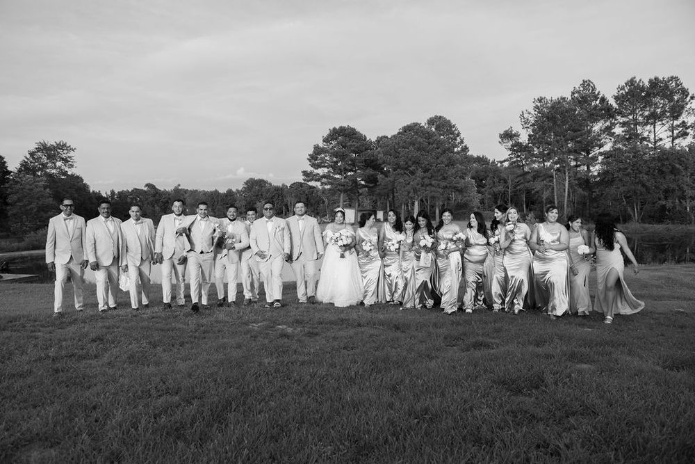 Black-and-white of the full wedding party, groomsmen in light suits and bridesmaids in satin gowns, crossing a field
