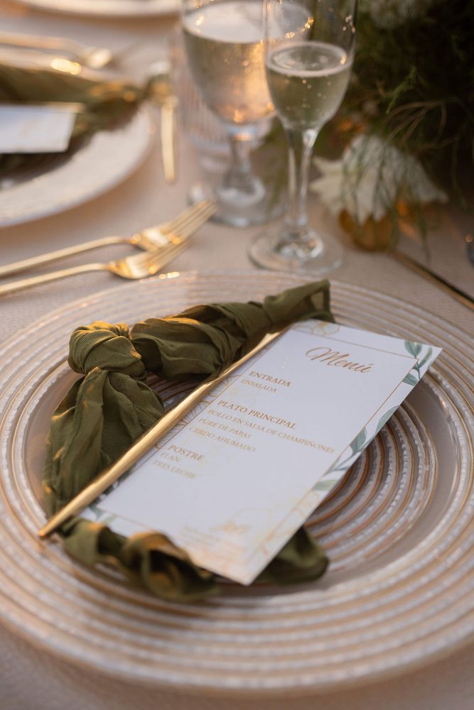 Dinner menu tucked into a knotted green napkin on a gold-rimmed charger, champagne glasses glowing at sunset