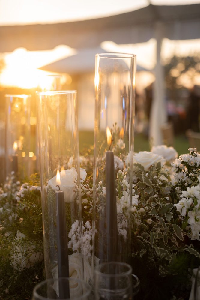 Dark taper candles in tall glass hurricanes among white roses and greenery, glowing in golden-hour light