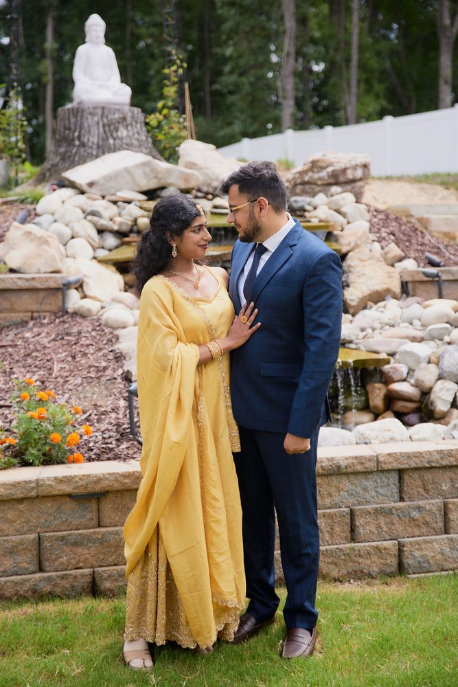 Couple in a yellow embroidered outfit and navy suit smiling at each other before a rock garden waterfall
