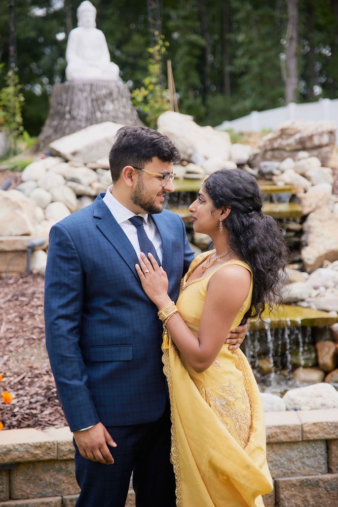 Bride in yellow with long curls resting her hand on the groom's chest beside a cascading stone fountain