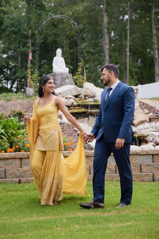 Couple walking hand in hand across the lawn, her yellow dupatta trailing, white statue and garden arch behind