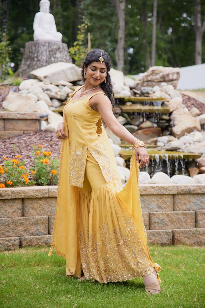 Bride twirling her yellow embroidered sharara and dupatta in front of a stone waterfall and marigolds