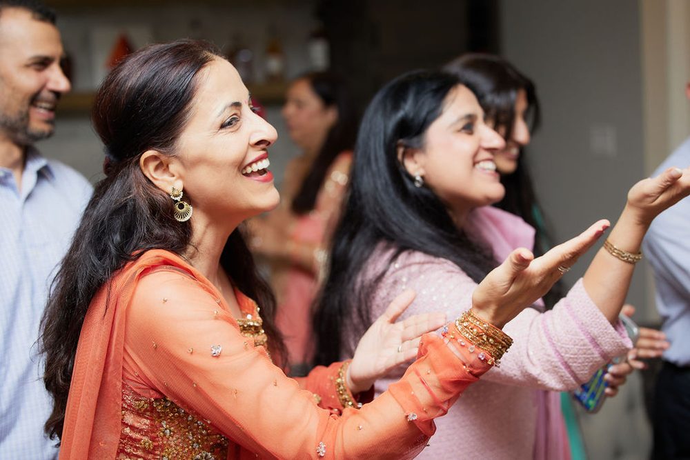 Laughing guests in an orange embroidered outfit and pink sweater clapping and dancing at the celebration