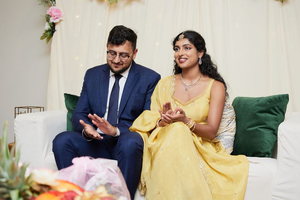 Couple seated on a white sofa with green pillows clapping along, draped backdrop with fairy lights behind them