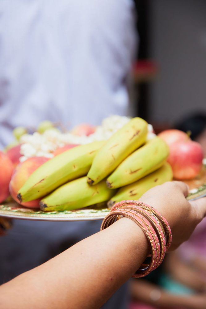 Hand wearing pink bangles carrying a platter of bananas, apples and white flowers