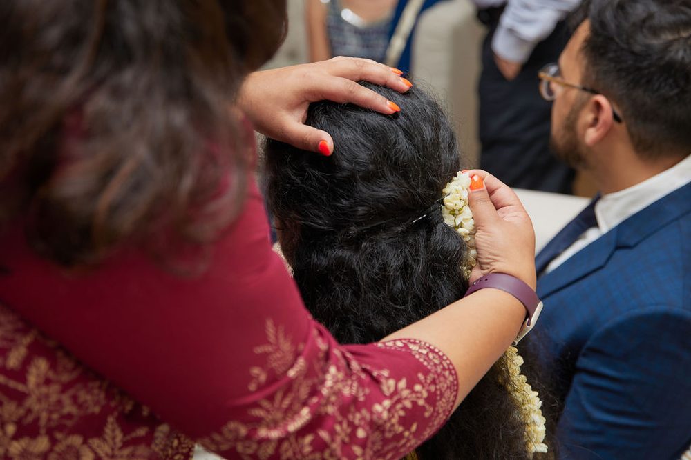 Relative in a maroon embroidered outfit pinning a jasmine garland into the bride's dark curls as the groom sits beside