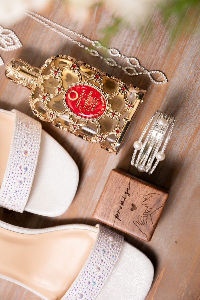 Gold perfume bottle with red label beside sparkly sandals, pearl cuff, crystal necklace and a wooden 'promise' ring box