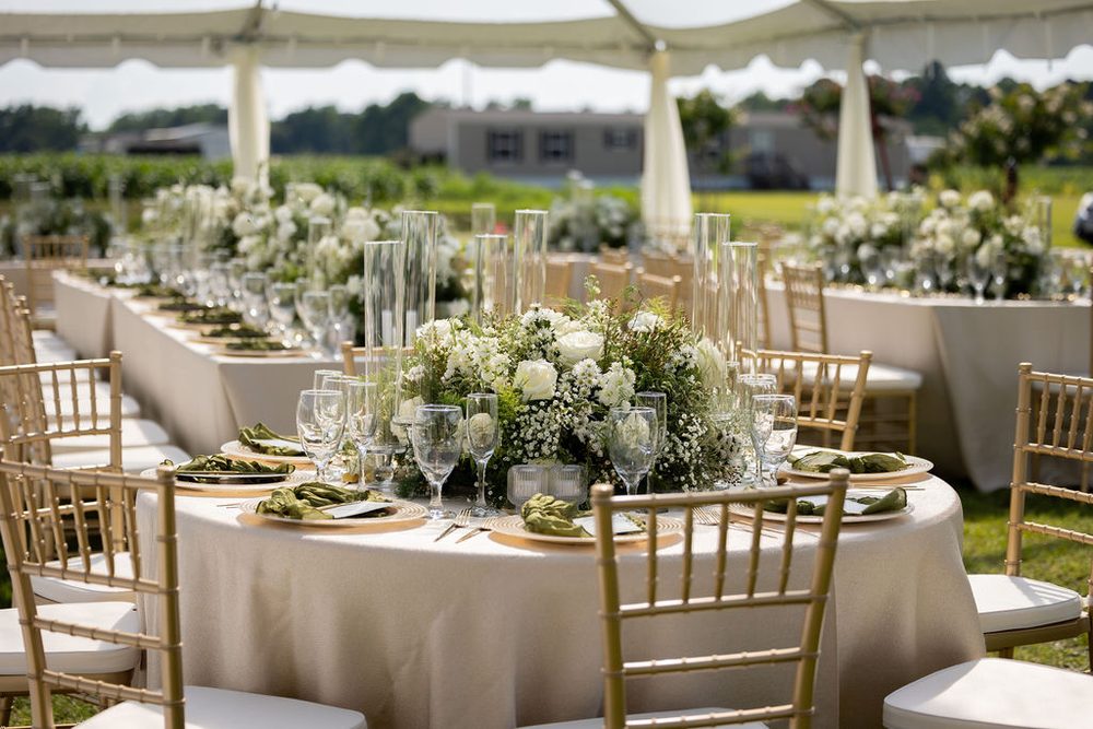 Round reception table under a white tent with gold chiavari chairs, green napkins and a white floral centerpiece