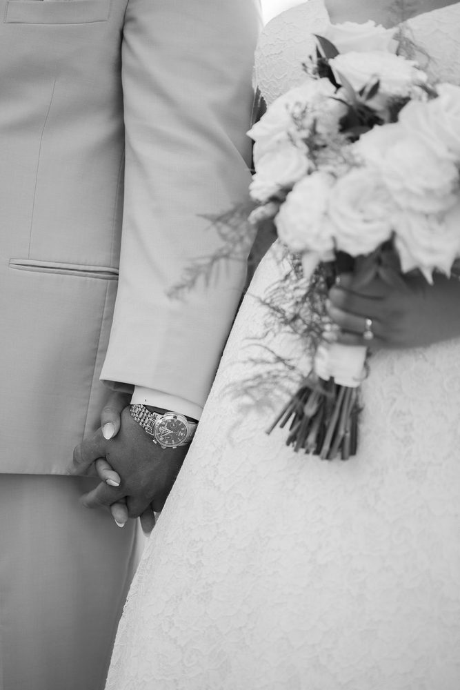 Black-and-white detail of the couple holding hands, his wristwatch beside her lace gown and rose bouquet