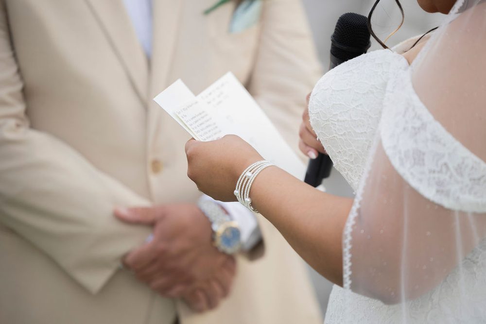 Bride in lace and pearl bracelets reading handwritten vows into a microphone beside the groom in a tan suit