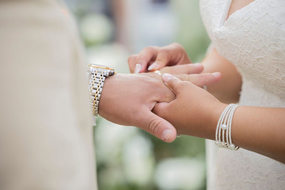 Close-up of the bride in a lace gown and pearl bracelet slipping a ring onto the groom's finger