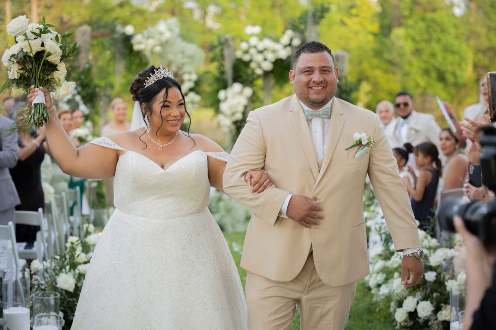 Joyful bride raising her white rose bouquet as she walks back up the aisle arm in arm with the groom