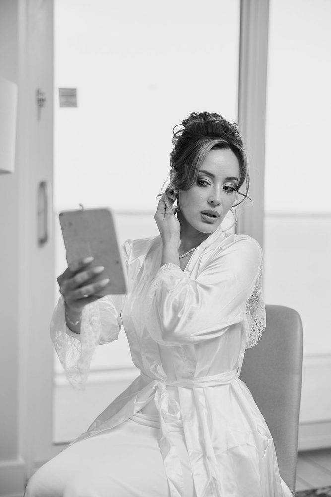 Black-and-white of the bride in her satin robe touching her earring while gazing into a hand mirror