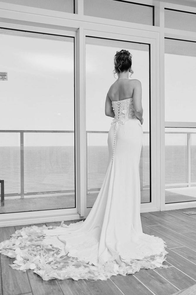 Black-and-white of the bride from behind in a corset-back gown with petal train, facing an ocean-view window