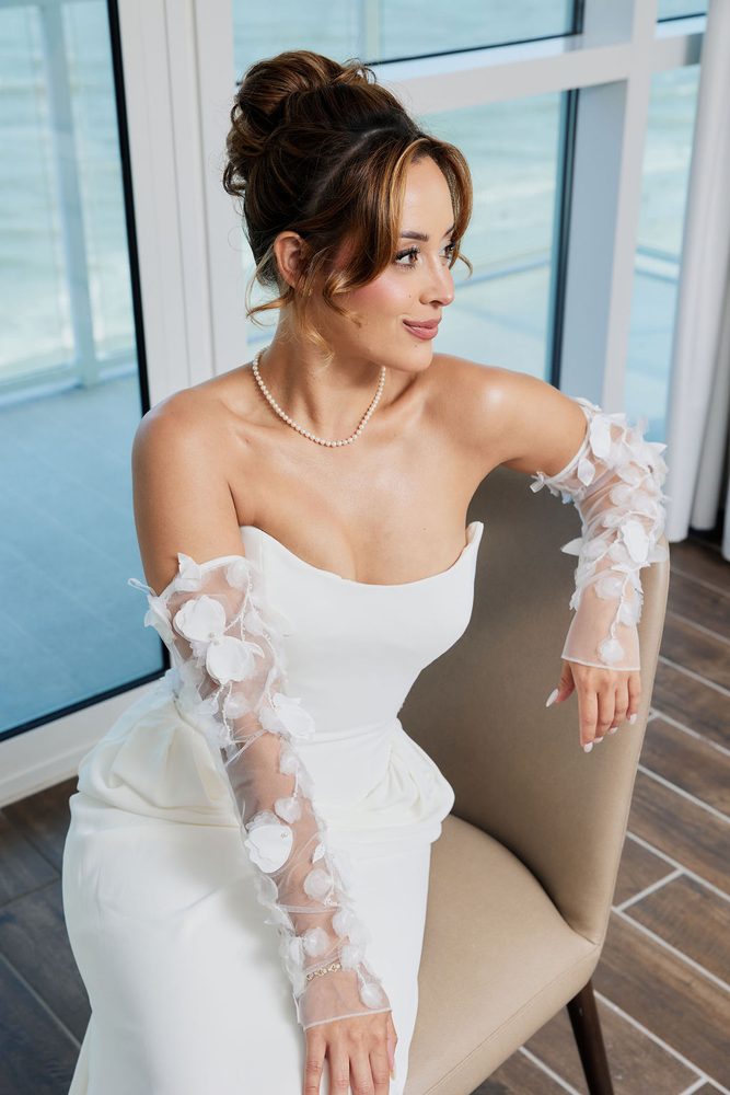 Bride seated in a strapless gown with sheer petal-appliqué sleeves and pearl necklace, smiling toward the window