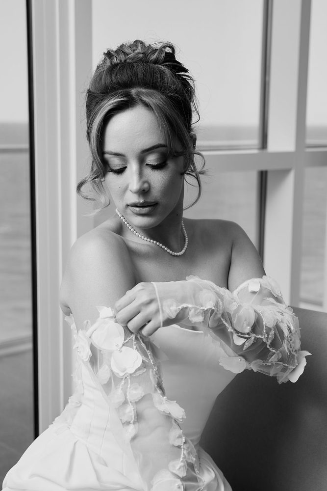 Black-and-white of the bride adjusting a sheer floral detachable sleeve, eyes lowered, pearls at her neck