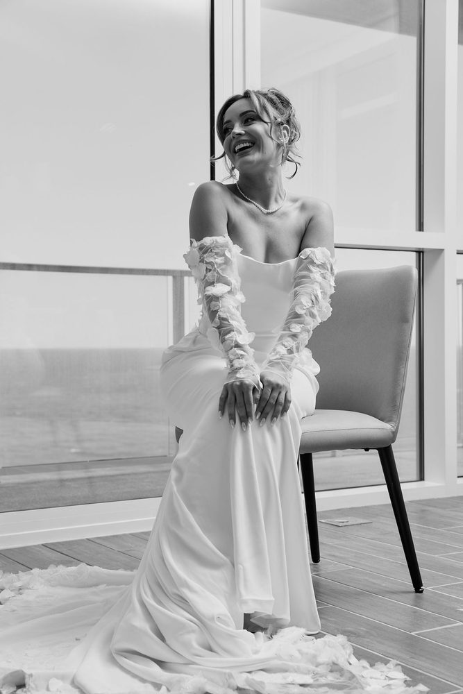 Black-and-white of the bride laughing on a chair in her strapless gown and petal sleeves, train pooled on the floor