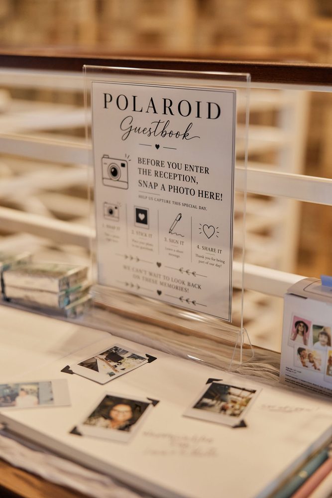 Polaroid guestbook sign on an acrylic stand above an album of instant photos at the reception entrance