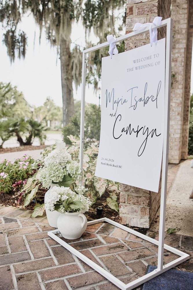 Wedding welcome sign on a white metal stand with hydrangeas in white pitchers by a brick entrance under Spanish moss