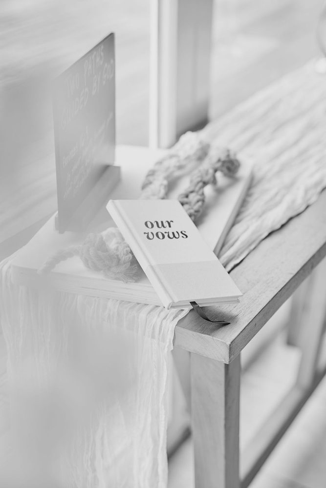 Black-and-white of an 'our vows' booklet and braided cord on a draped wooden ceremony table beside a sign