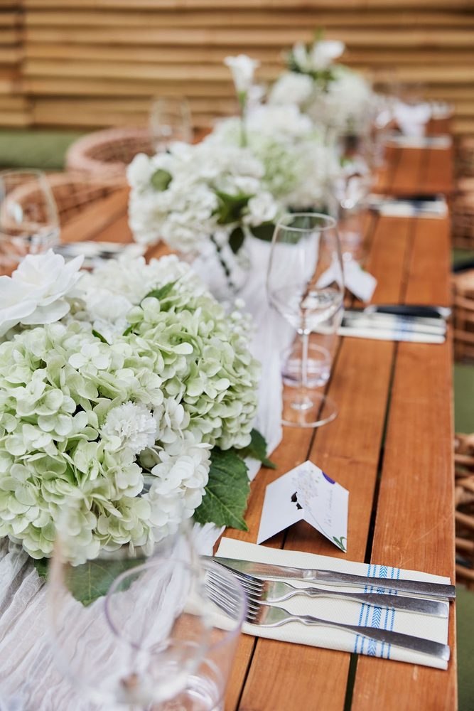 Long wooden table set with a green and white hydrangea runner, wine glasses, striped napkins and place cards