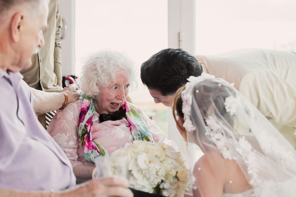 Elderly guest in a floral scarf beaming in delight as the bride and groom lean in to greet her