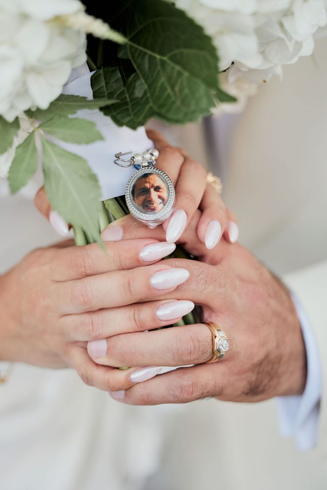 Couple's hands clasped around the bouquet stems, a photo charm of a loved one pinned to the ribbon wrap