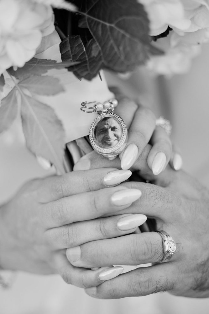 Black-and-white close-up of the photo locket of a loved one on the bouquet, the groom's signet ring and the bride's nails