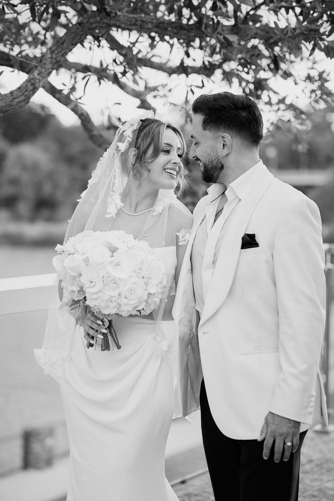 Black-and-white candid of the bride laughing up at the groom beneath tree branches, holding a rose bouquet