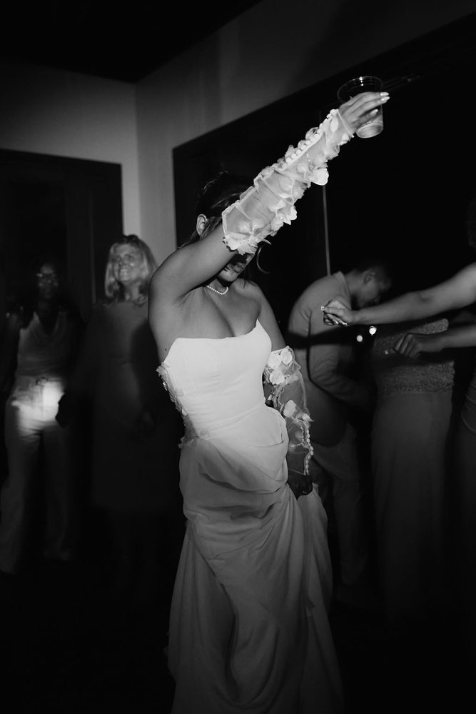 Black-and-white of the bride dancing at the reception, arm raised with a cup, sheer petal sleeve lit by the flash