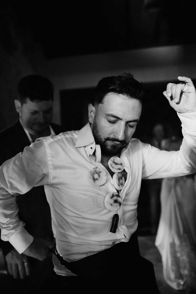 Black-and-white of the groom dancing in his white shirt with a playful party necklace, one arm raised