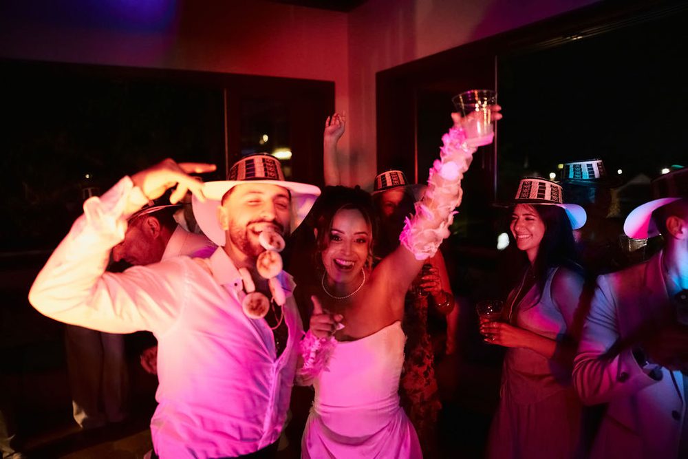 Bride and groom dancing under pink party lights with guests in striped wide-brim hats, cups raised high