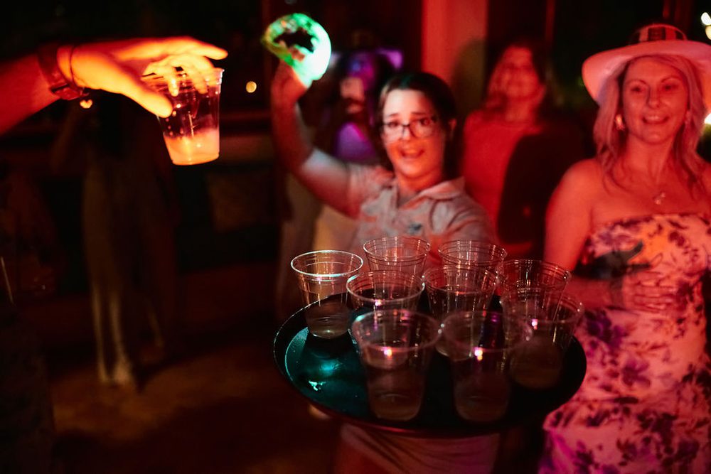 Tray of drinks in plastic cups carried across the dance floor as guests in party hats celebrate under red lights