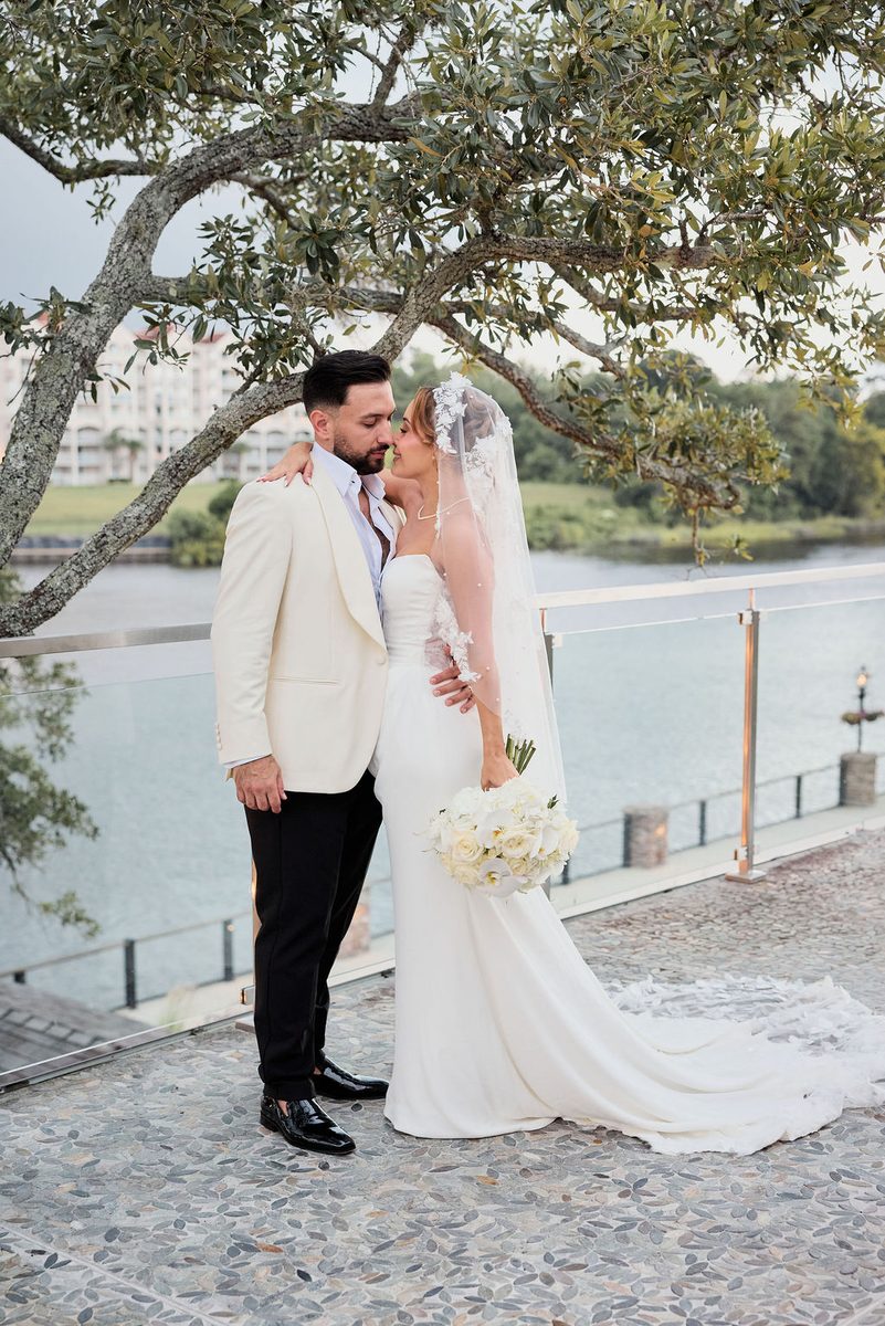 Wedding photography: bride and groom nuzzling under an oak tree on a waterfront terrace, white rose bouquet and lace veil
